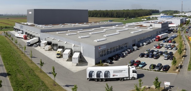Luftaufnahme einer Lagerhalle mit Verladerampen, mehreren Lastwagen und einem Sattelzug auf der Zufahrt.