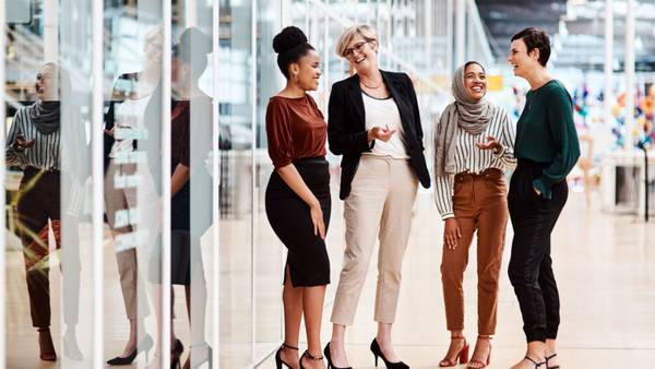 Five businesswomen talking in a bright office lobby behind a glass wall.