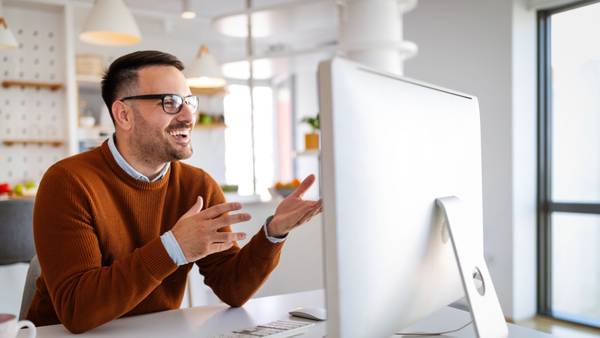 Man in an orange jumper gesturing during a video call in front of a monitor.