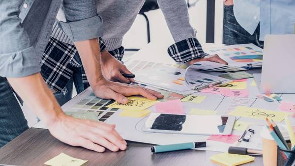 Three people standing over a table covered in sketches, sticky notes and colour samples.