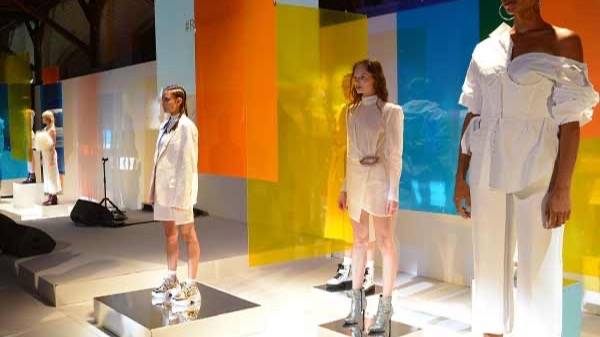 Models in white outfits on plinths in front of coloured acrylic panels at a fashion presentation.