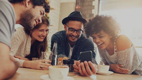 Four young people at a cafe table laughing at a smartphone together.