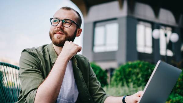 Bearded man with glasses sitting outdoors with a laptop in front of a modern building.