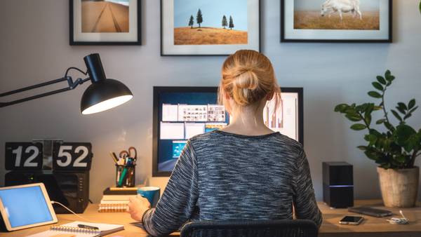 Woman seen from behind at a desk with two monitors, a desk lamp and framed pictures on the wall.